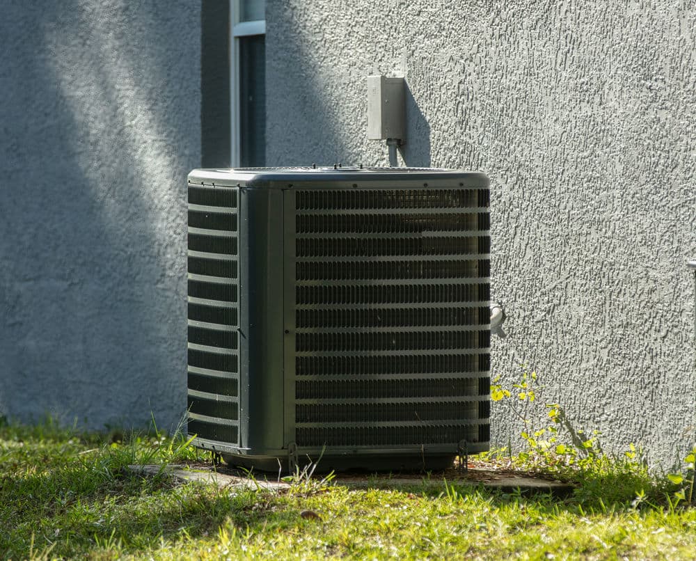 Air conditioning unit installed outside a house on a grassy patch near a textured wall.