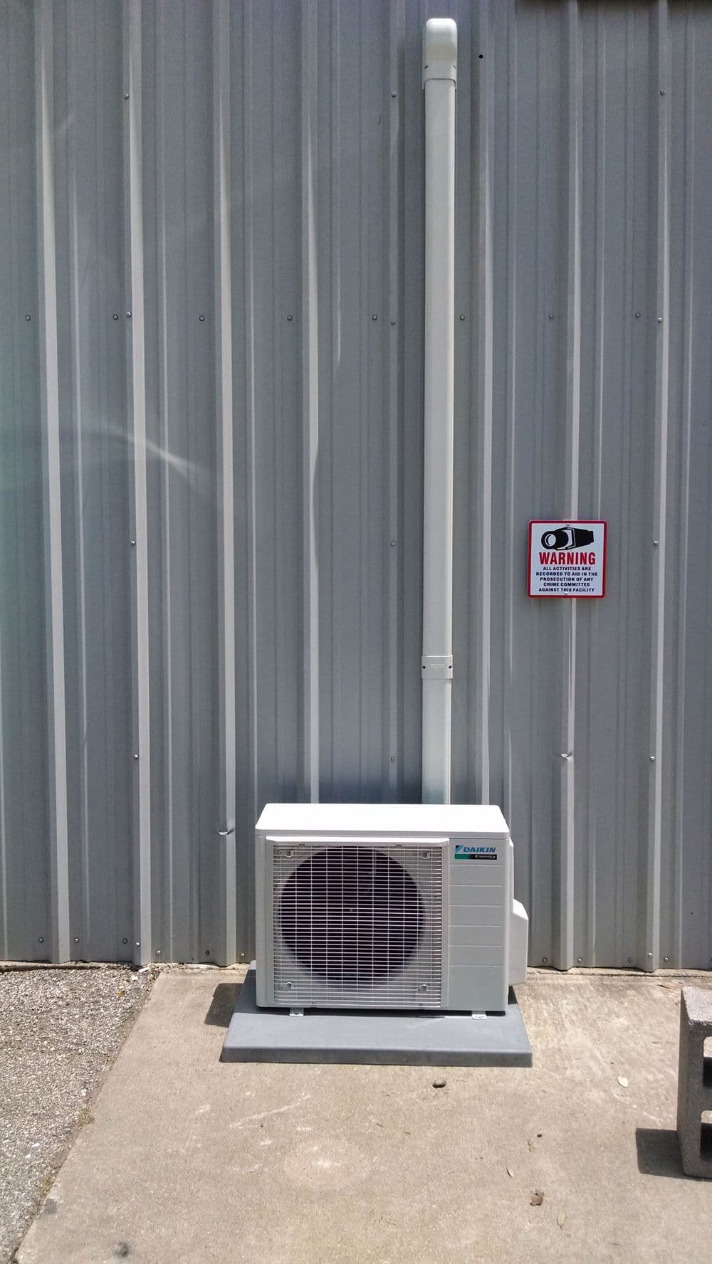 Air conditioning unit installed on a concrete surface beside a metal wall, with warning sign.