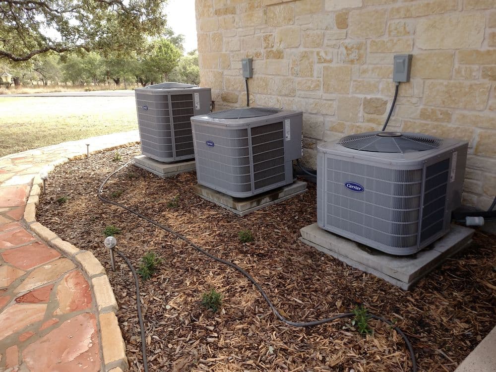 Three outdoor air conditioning units next to a stone wall, with landscaping and electrical connections.
