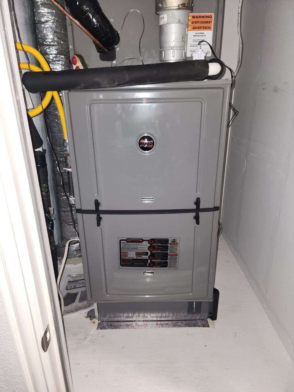 Gray furnace located in a utility closet with safety warning signs and plumbing connections.