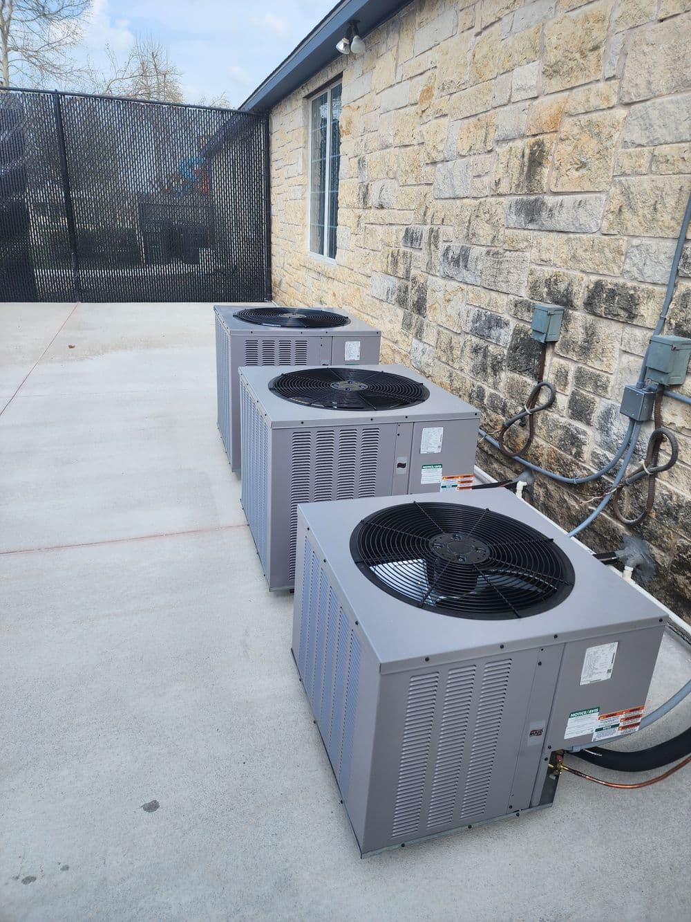 Three outdoor air conditioning units beside a stone building on a concrete surface.