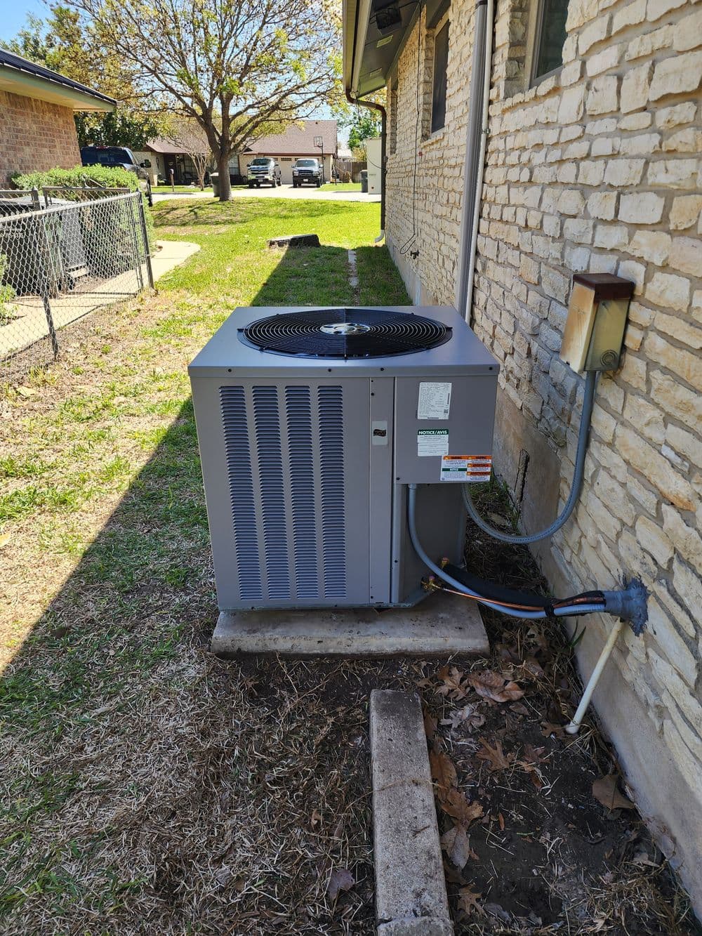 Air conditioning unit installed beside a house, surrounded by grass and trees.