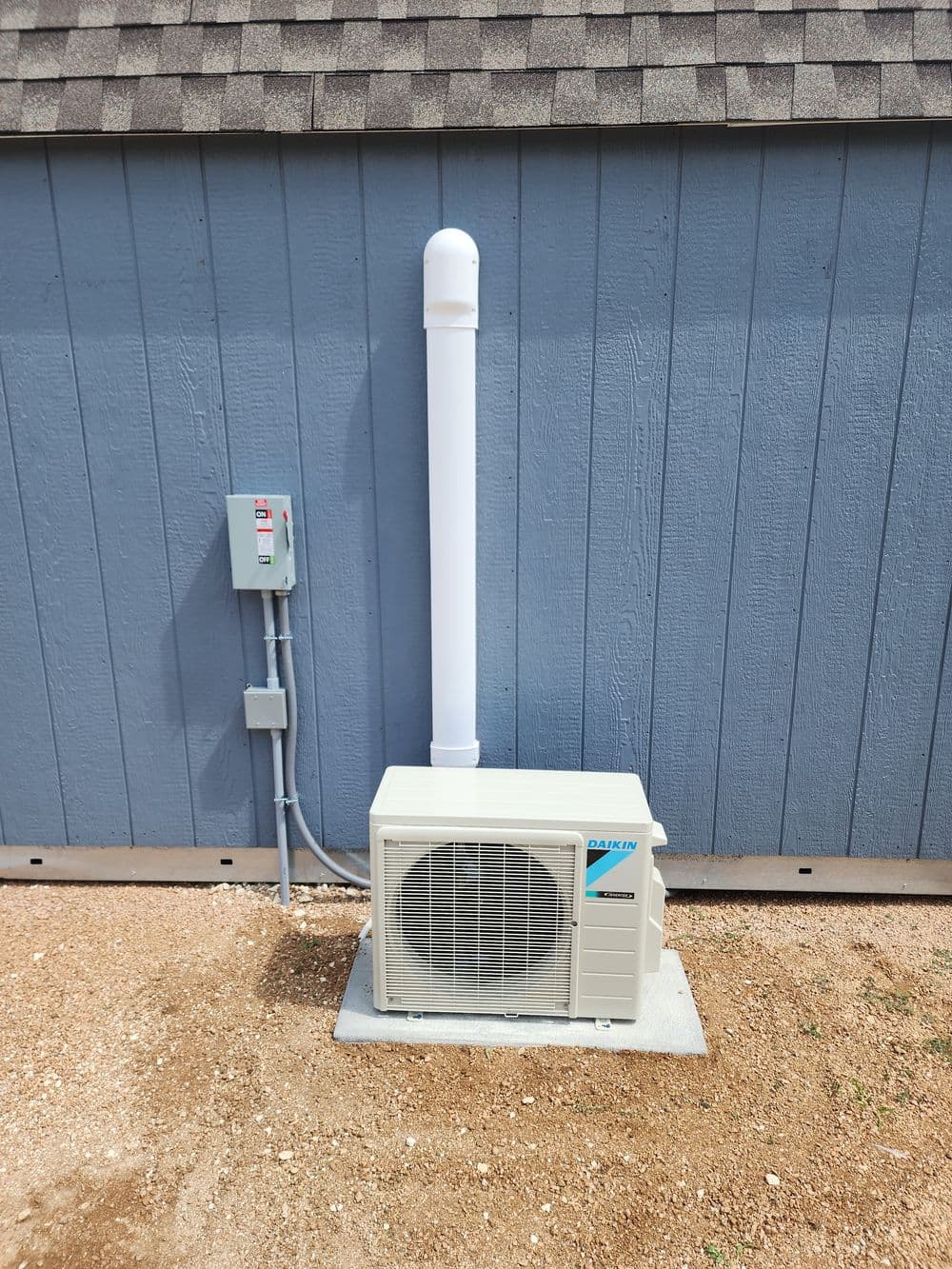 Air conditioning unit installed beside a blue shed, with a white exhaust pipe and electrical panel.